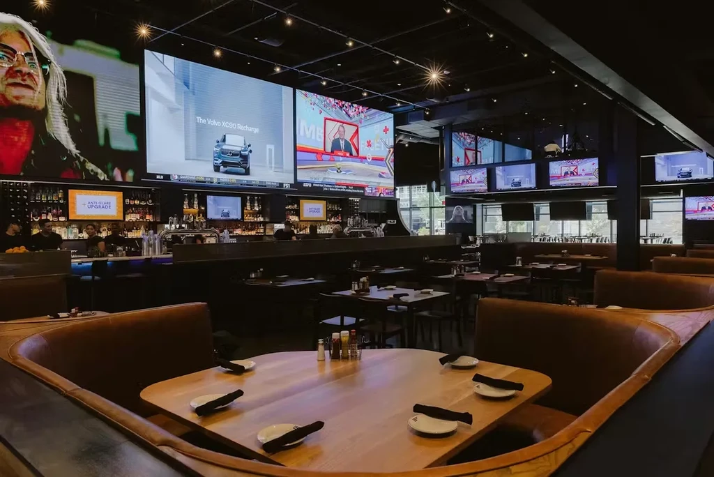 Modern sports bar with dining booths, an illuminated bar, and multiple screens.