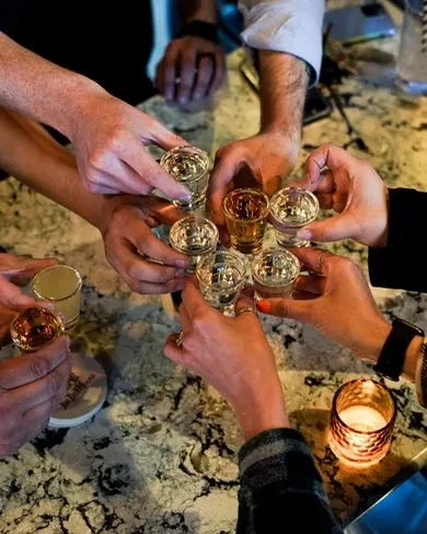 Multiple hands clinking shot glasses on a mottled countertop with a candle.