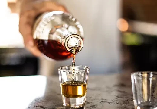 Hand pouring dark amber liquid into a shot glass.
