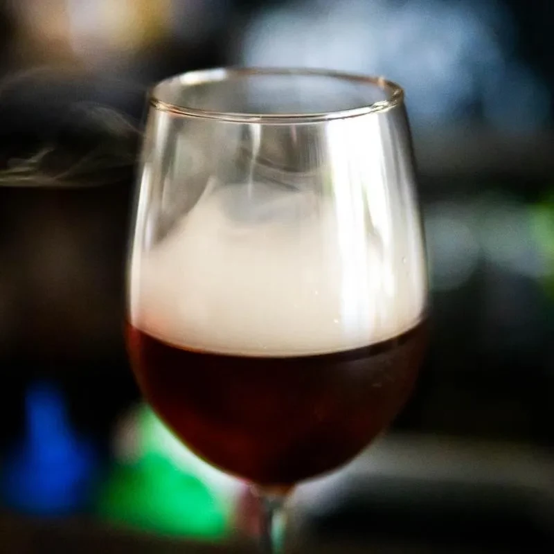 Wine glass with dark red cocktail and swirling white smoke.
