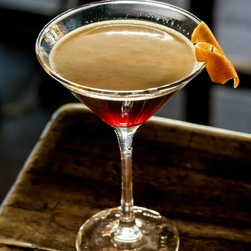 Layered cocktail in martini glass with red liquid, tan foam, orange peel.