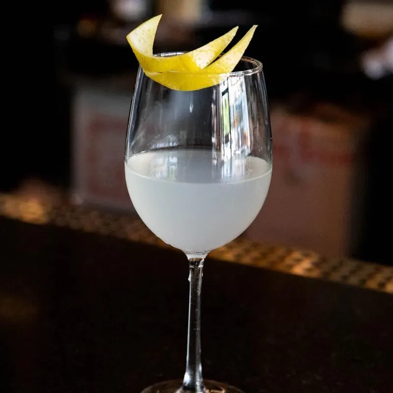 Pale cocktail in wine glass, garnished with bright yellow lemon peel.