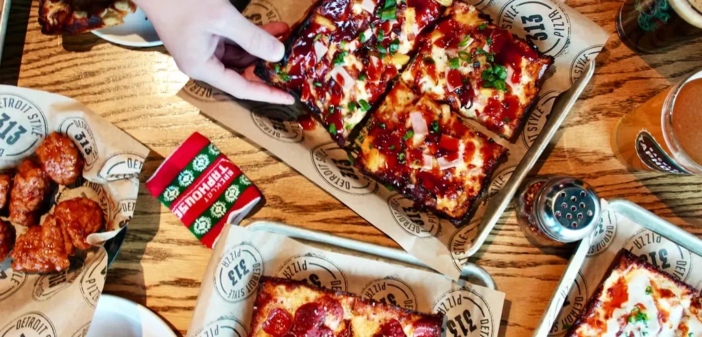 A hand takes Detroit-style pizza from a table with wings and beer.