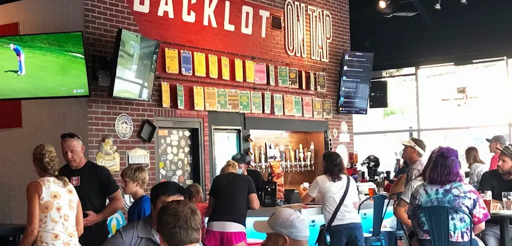 Busy brewpub bar with "BACKLOT ON TAP" sign, taps, and TVs.
