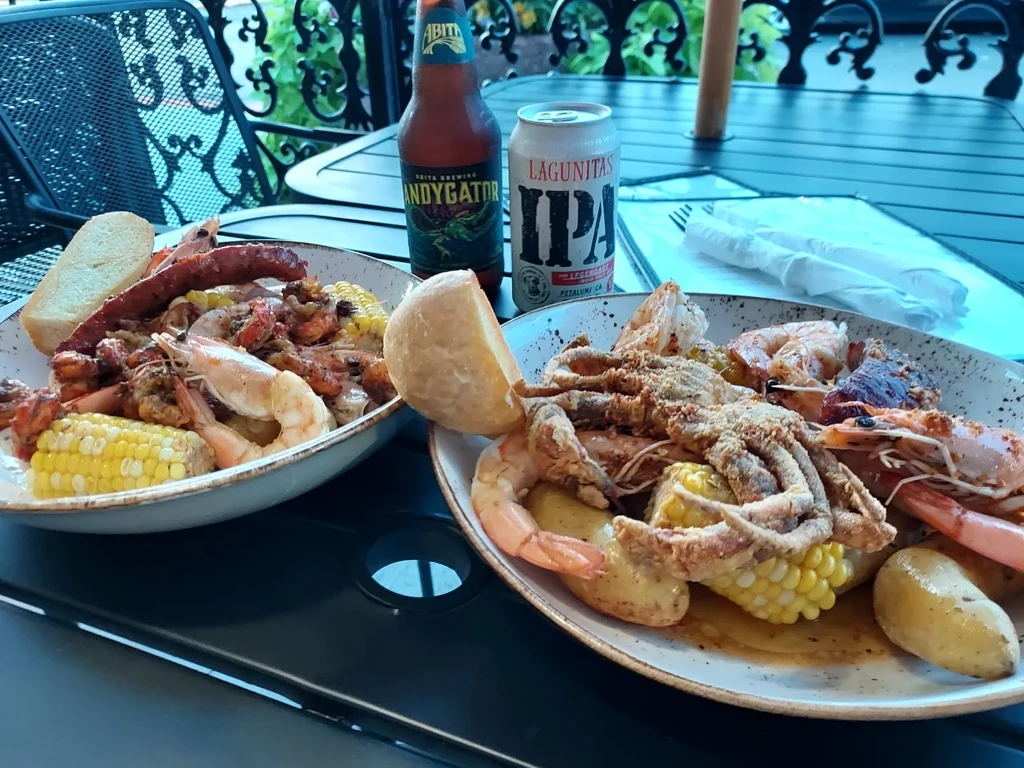 Two plates of outdoor seafood boil with shrimp, corn, sausage, beer.