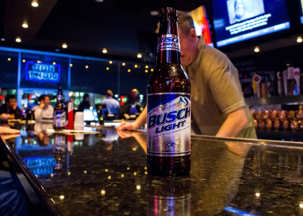 Focused Busch Light bottle on reflective bar in dim pub.