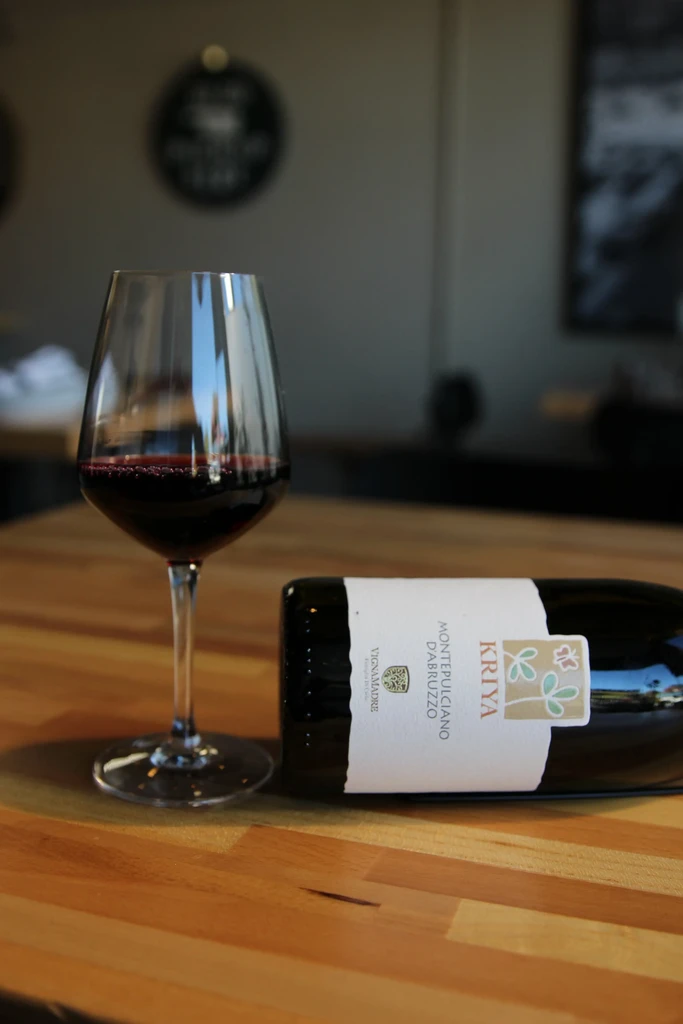 Red wine in glass with Kriya Montepulciano d'Abruzzo bottle on wooden table.