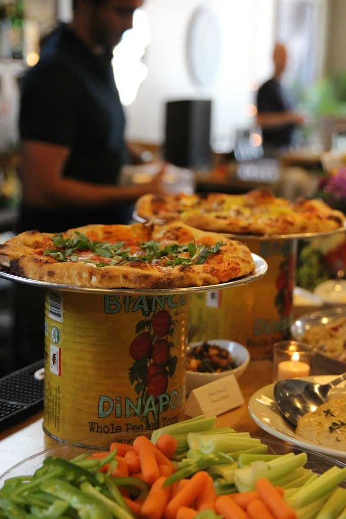 Buffet display: fresh vegetables, two pizzas elevated by tomato cans.