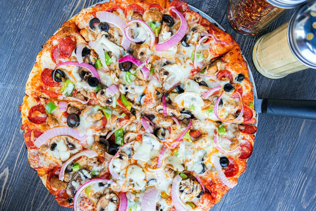 Loaded pizza with pepperoni, vegetables, cheese on a rustic wooden table.