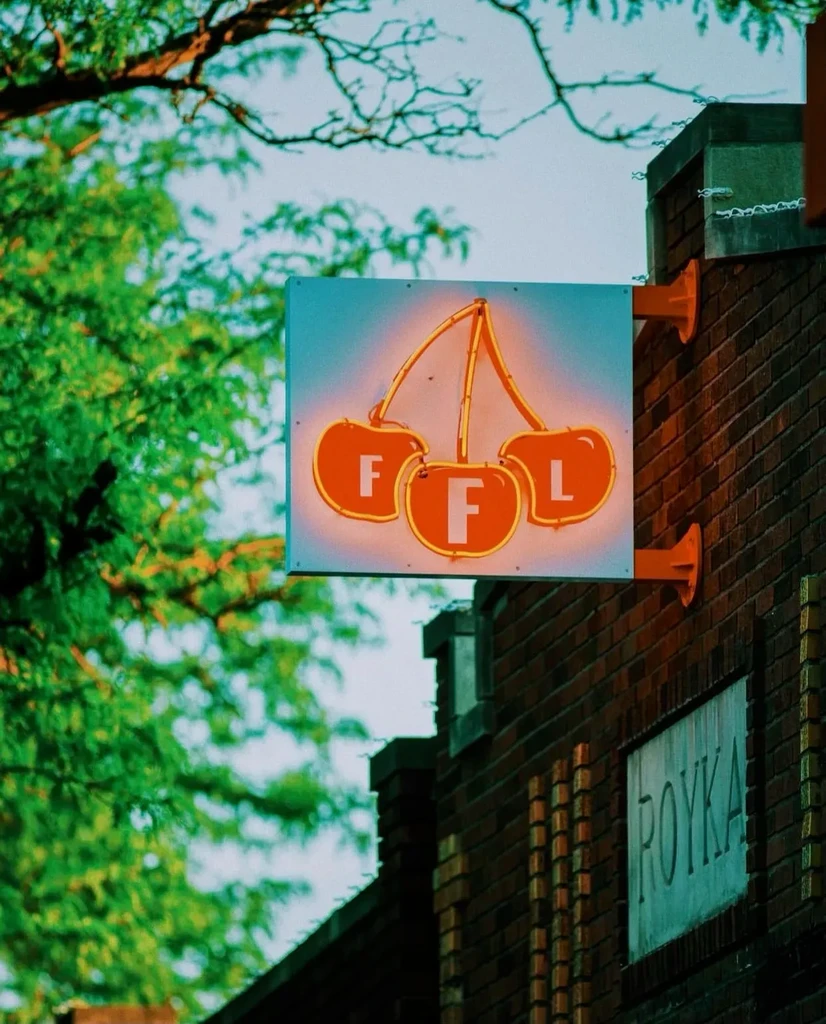 Orange-red neon cherry 'FFL' sign on brick wall above 'ROYKA' plaque.