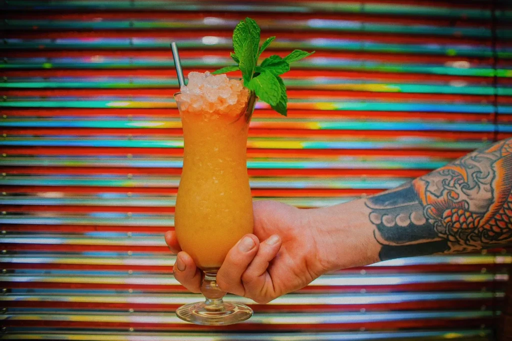 Tattooed arm holding an orange frozen drink with mint and straw.