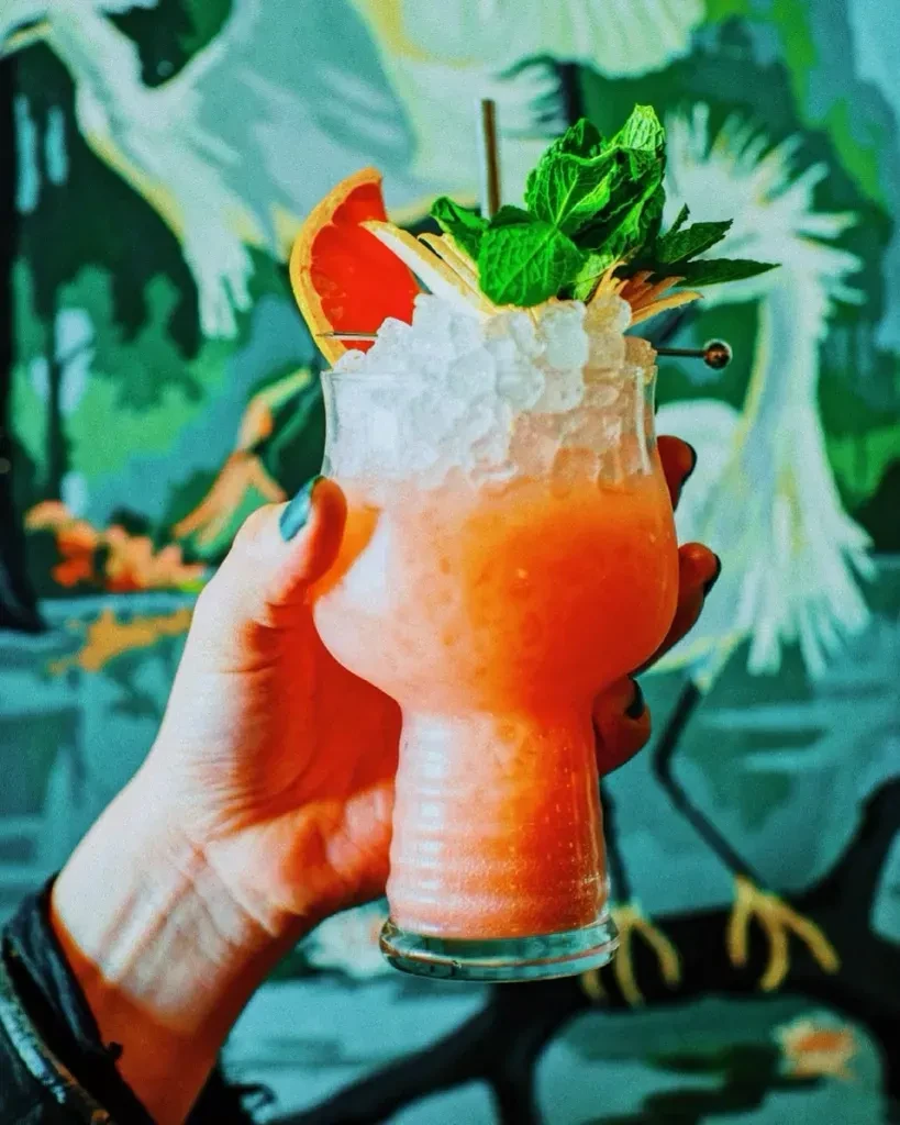 Hand holding orange-pink cocktail with grapefruit, mint, against a teal mural.