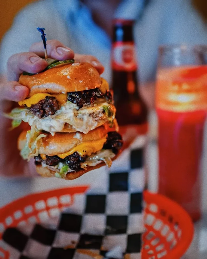 Hand holds a multi-patty cheeseburger with cheese, lettuce, and pickle.