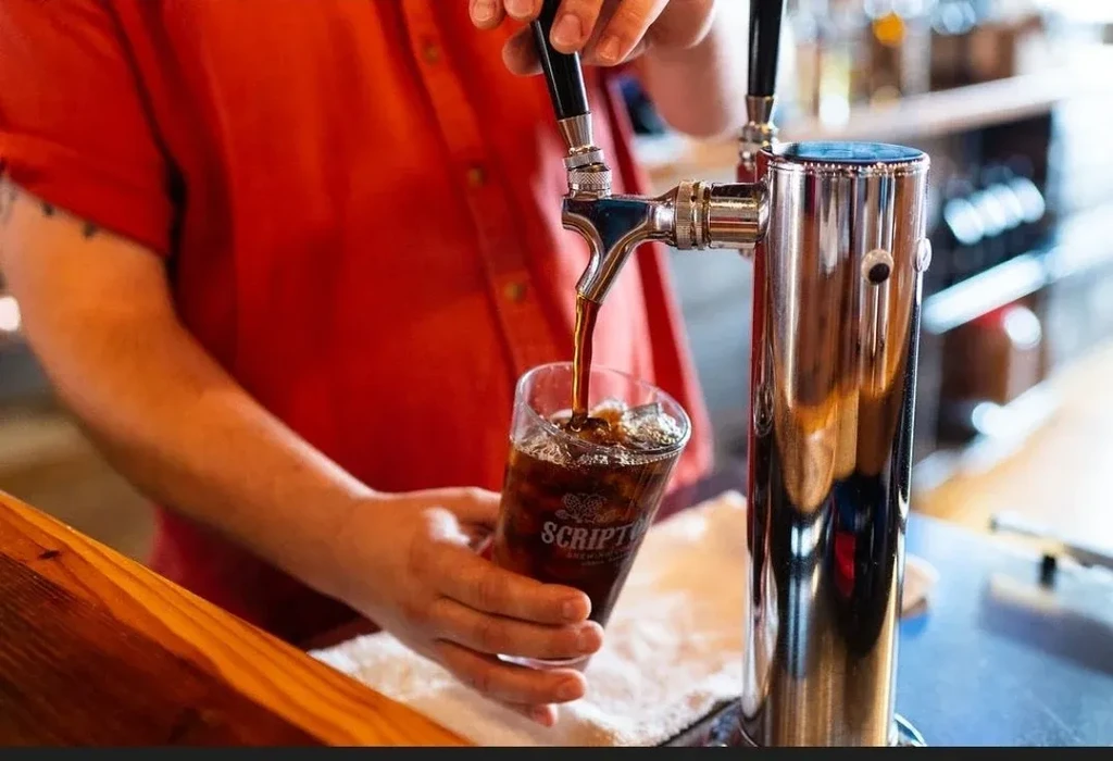 Hands filling Scripto glass with dark iced beverage from tap.