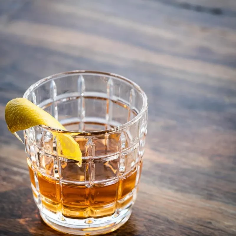 Close-up: amber drink in textured glass, lemon peel, dark wood.
