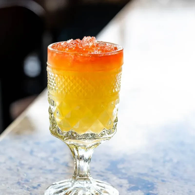 Orange-yellow layered cocktail with crushed ice in a decorative glass.