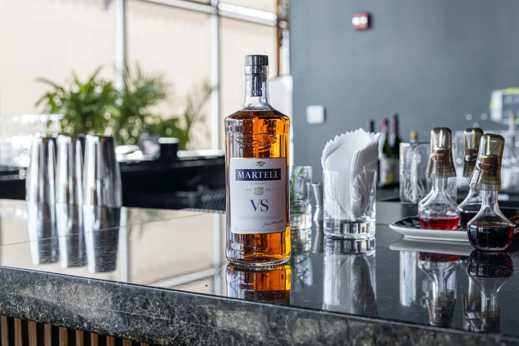 Martell Cognac VS bottle on a dark bar counter with bar equipment.
