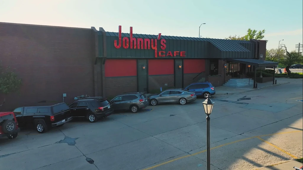 Daytime exterior of Johnny's Cafe, brick facade, red neon sign, parked cars.