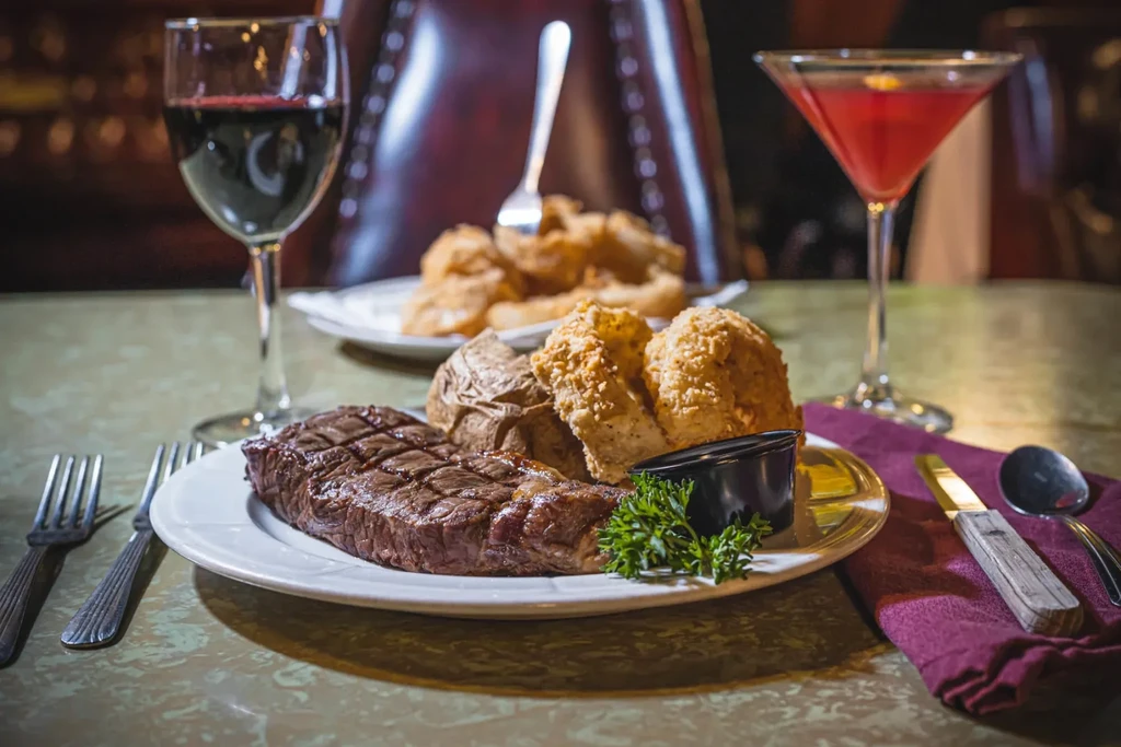 Grilled steak, baked potato, biscuits, red wine, and cocktail on a table.