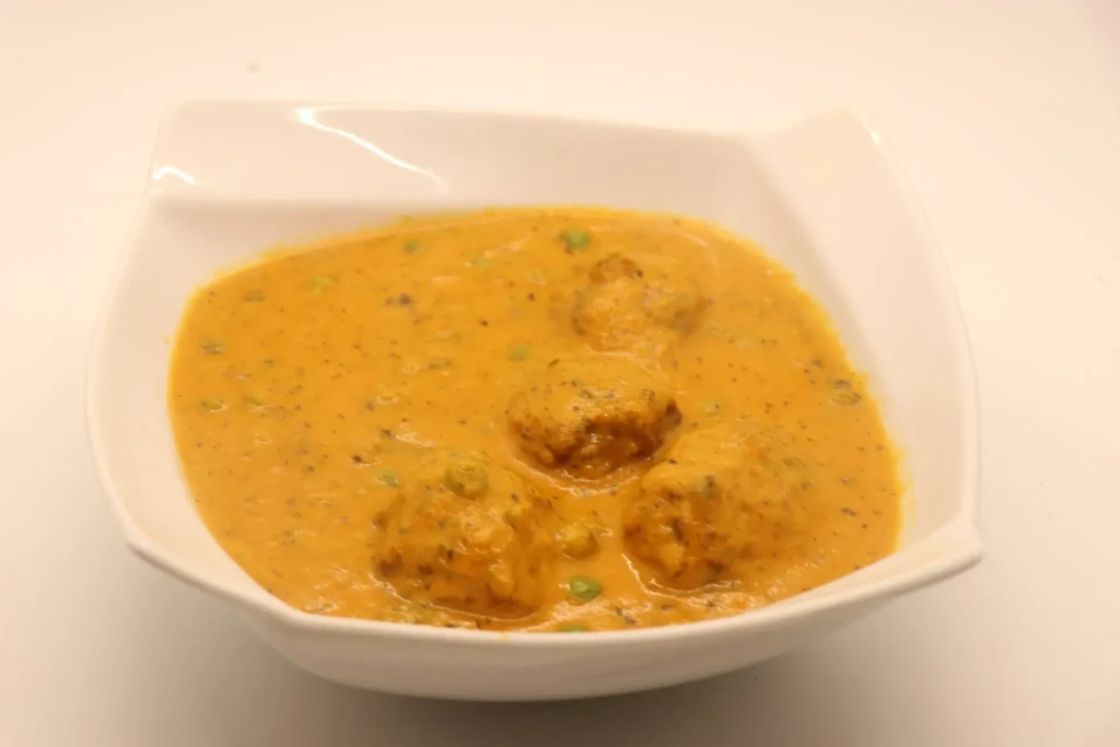 Creamy orange kofta curry with green peas in a white bowl.