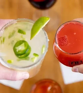 Two hands hold a green cocktail with jalapeño and red drink.