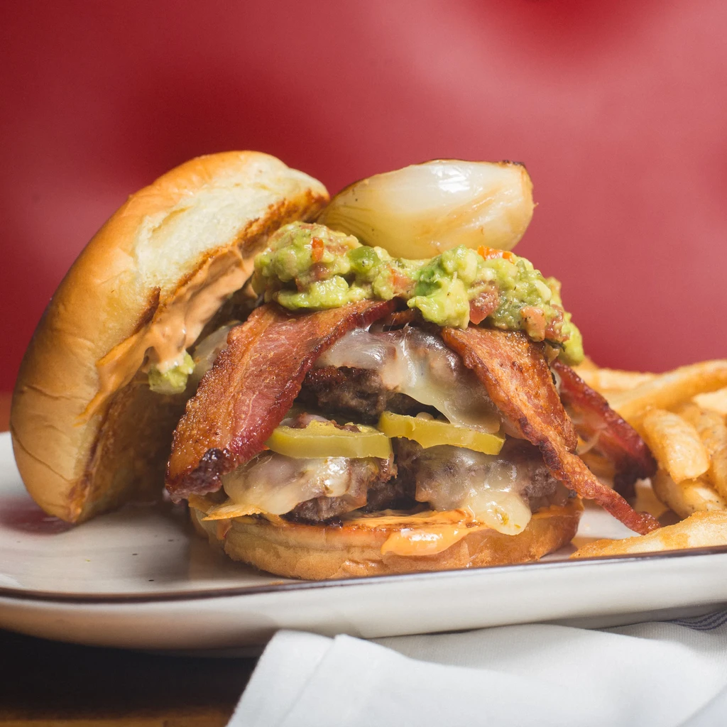 Close-up burger with bacon, cheese, guacamole, onion, fries, red background.