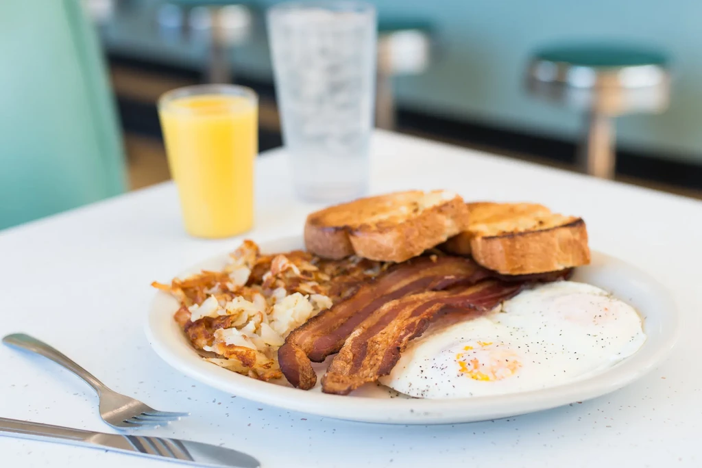 A plate of fried eggs, bacon, hash browns, toast, juice, and water.