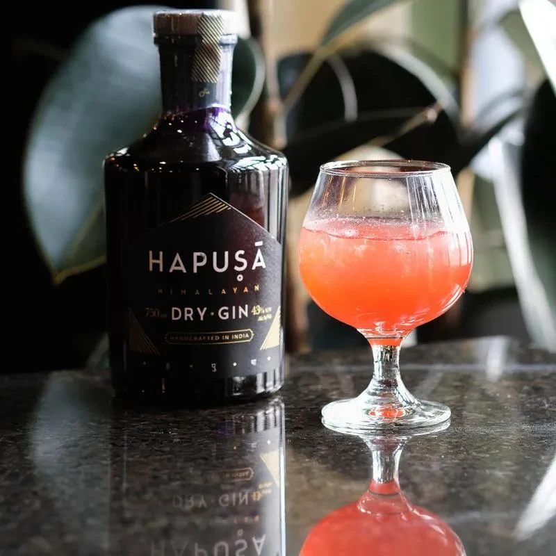 Hapusa Gin bottle and reddish cocktail on a reflective surface.
