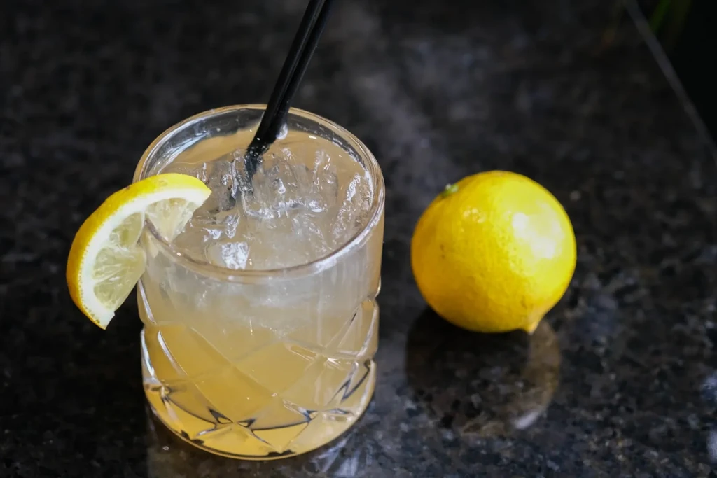 Iced yellow drink with lemon slice, whole lemon, and black straw.