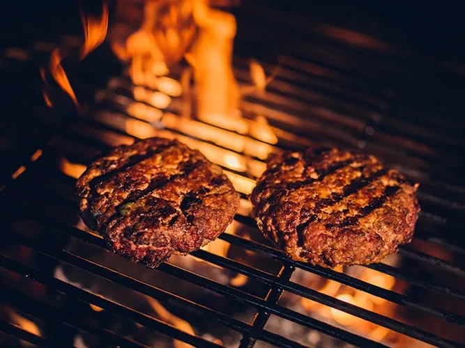 Two hamburger patties grilling over open flames on a metal grate.
