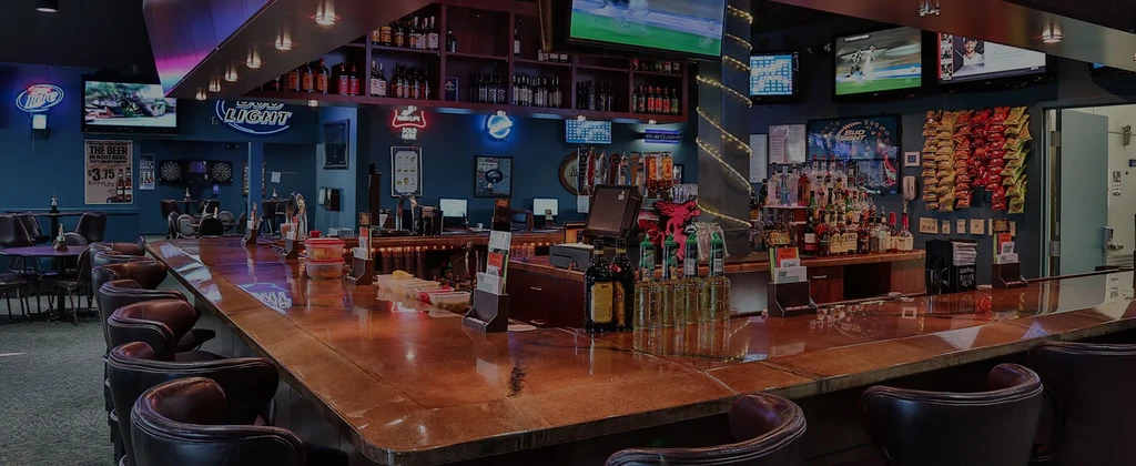 Empty sports bar interior with L-shaped counter, stocked bar, TVs, and neon signs.