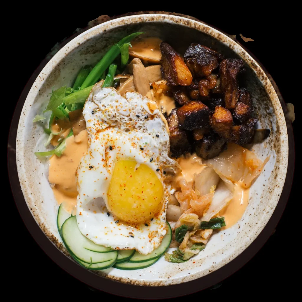 Top-down: rustic bowl with sunny egg, glazed meat, colorful vegetables, sauce.