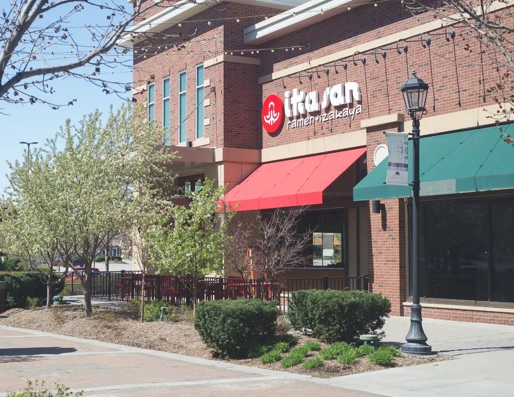 Ikasan ramen restaurant exterior, red awning, blooming trees, outdoor patio.