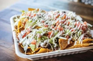 Large nachos in a white container, with lettuce, tomatoes, creamy sauce.