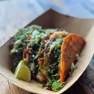 Three birria-style tacos with cilantro and green sauce in a paper container.