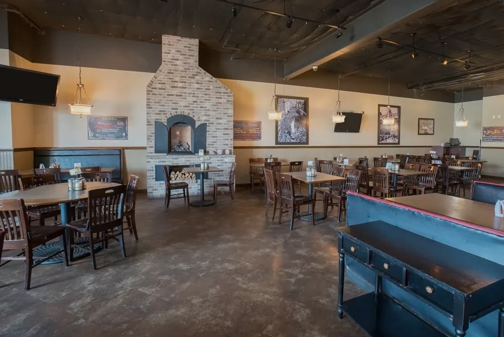 Empty restaurant with brick fireplace, wooden tables, and an industrial ceiling.