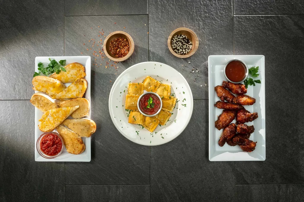Garlic bread, fried ravioli, and chicken wings with sauces on dark tile.