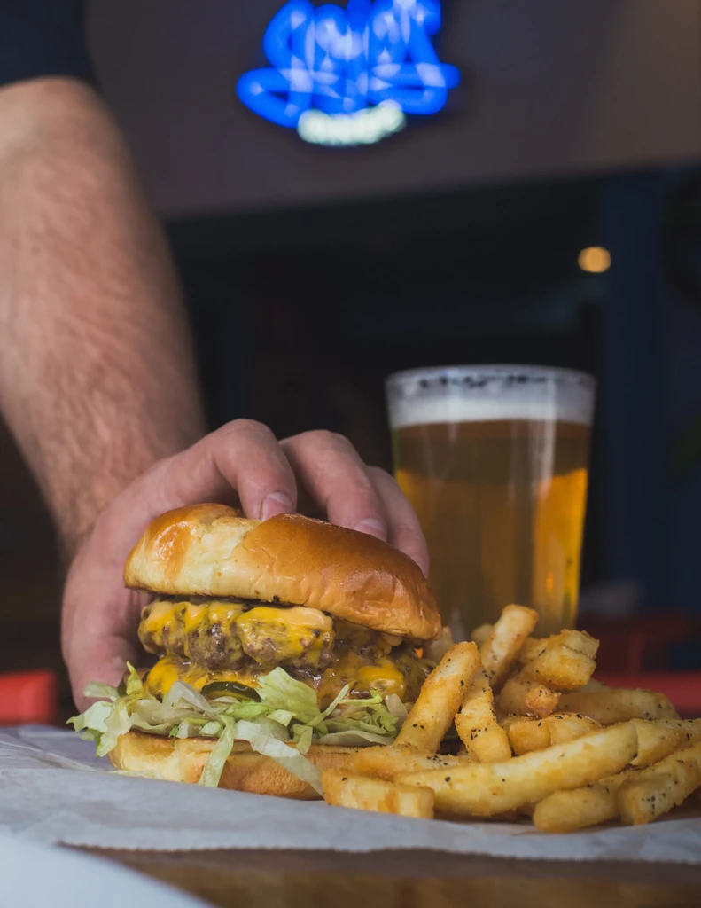 Hand holds double cheeseburger with jalapeños, fries, blurred beer, neon.