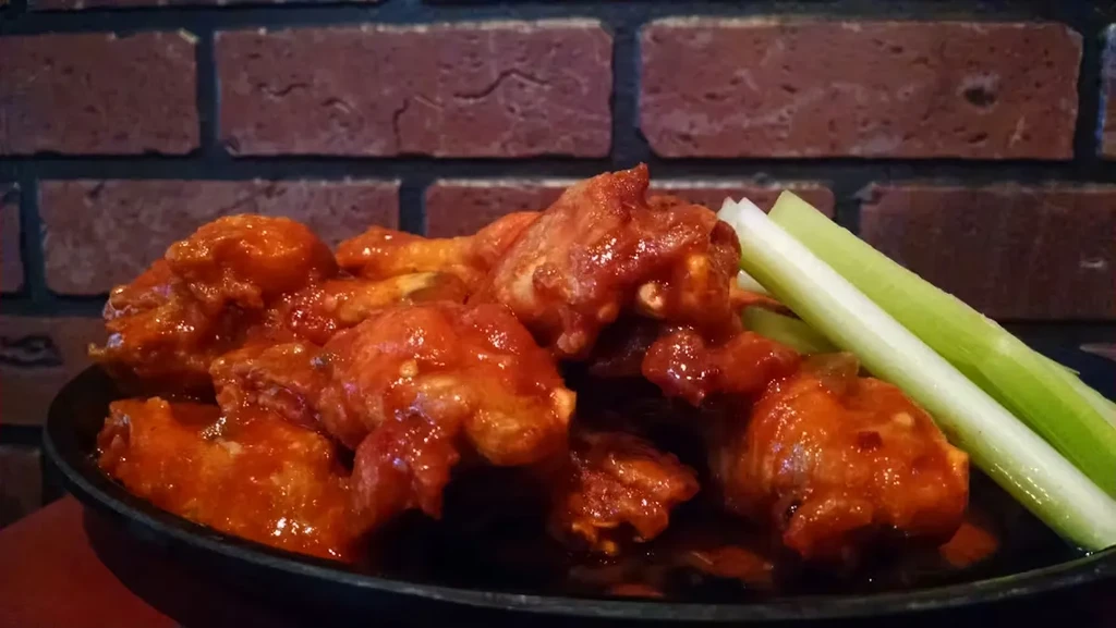 Orange sauced chicken wings and celery against a brick wall.