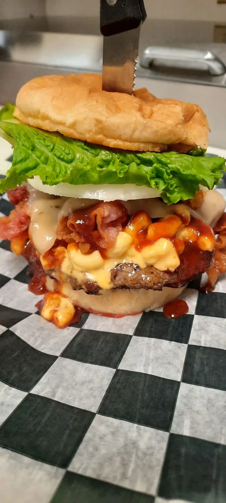Giant mac and cheese burger with bacon, BBQ sauce, held by knife.