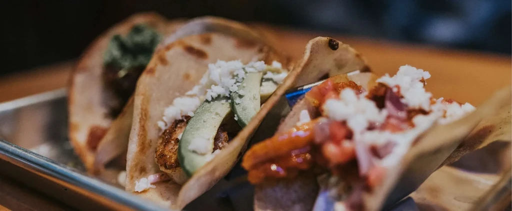 Three tacos in a tray with avocado, protein, cheese, and salsa.