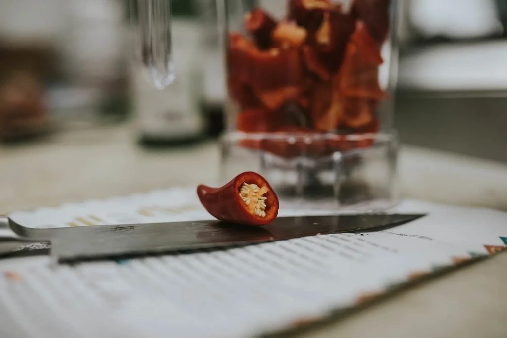 Halved red chili pepper on a silver knife, with chopped peppers nearby.