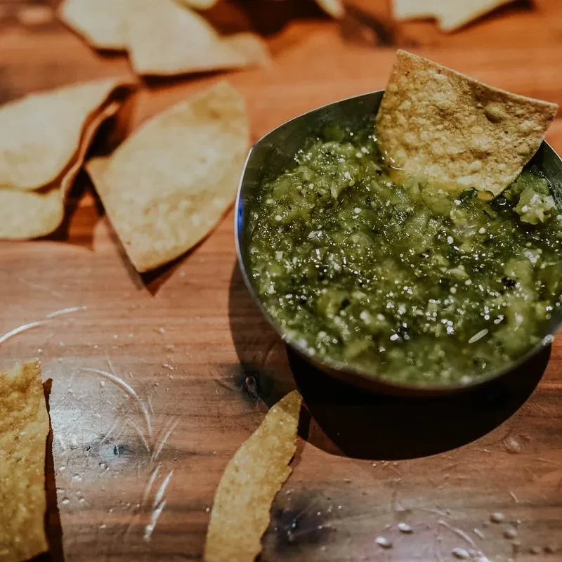 Metallic bowl of chunky green salsa and tortilla chips on wood.
