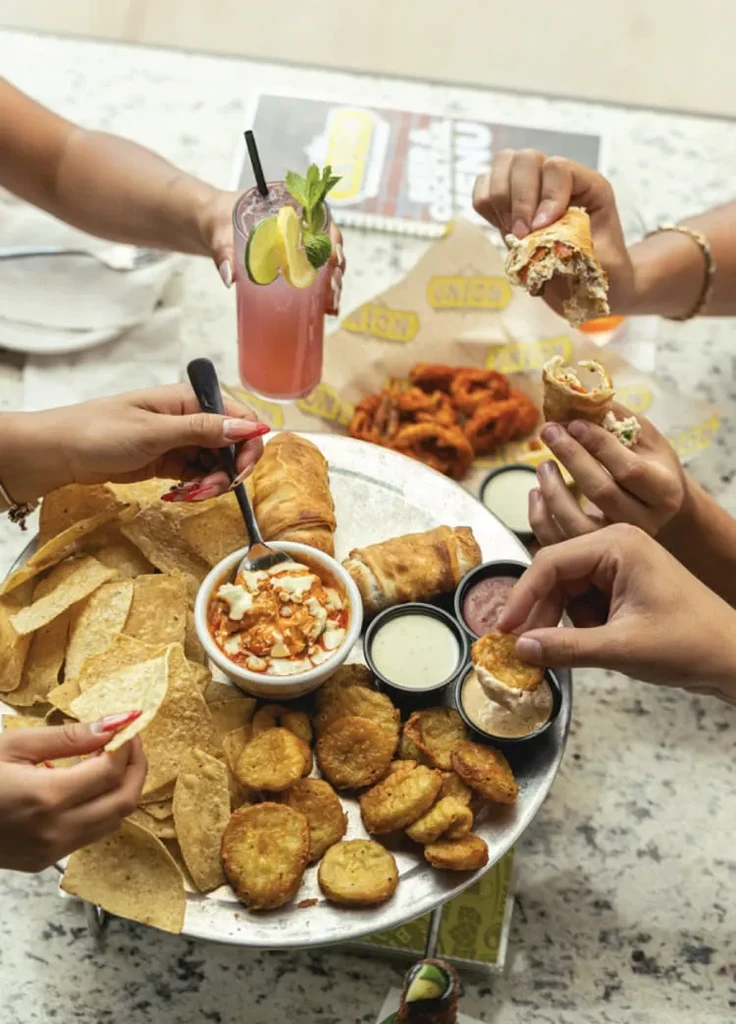 Hands share fried appetizers, chips, and a cocktail from a large tray.