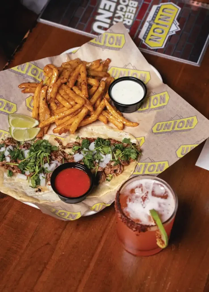 Overhead shot of street tacos, fries, sauces, and a garnished cocktail.