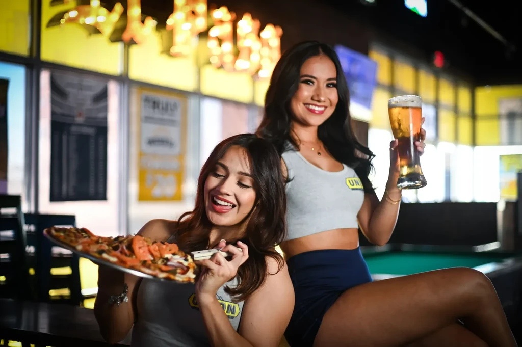 Two smiling women in a restaurant, one with pizza, one with beer.
