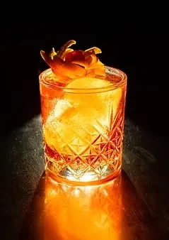 Amber cocktail in cut-glass tumbler, spherical ice, glowing orange peel.