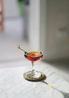Reddish-brown cocktail in coupe glass, cherry garnish, on woven coaster.