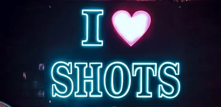 Vibrant blue and pink neon sign reads 'I ❤️ SHOTS'.