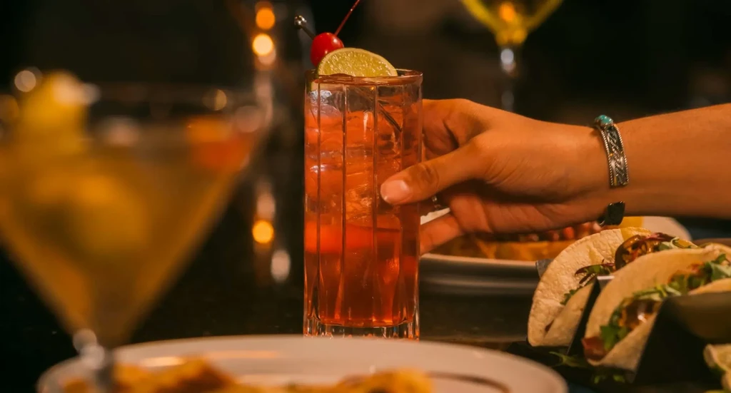 Hand holding garnished cocktail; blurred tacos and drinks in background.